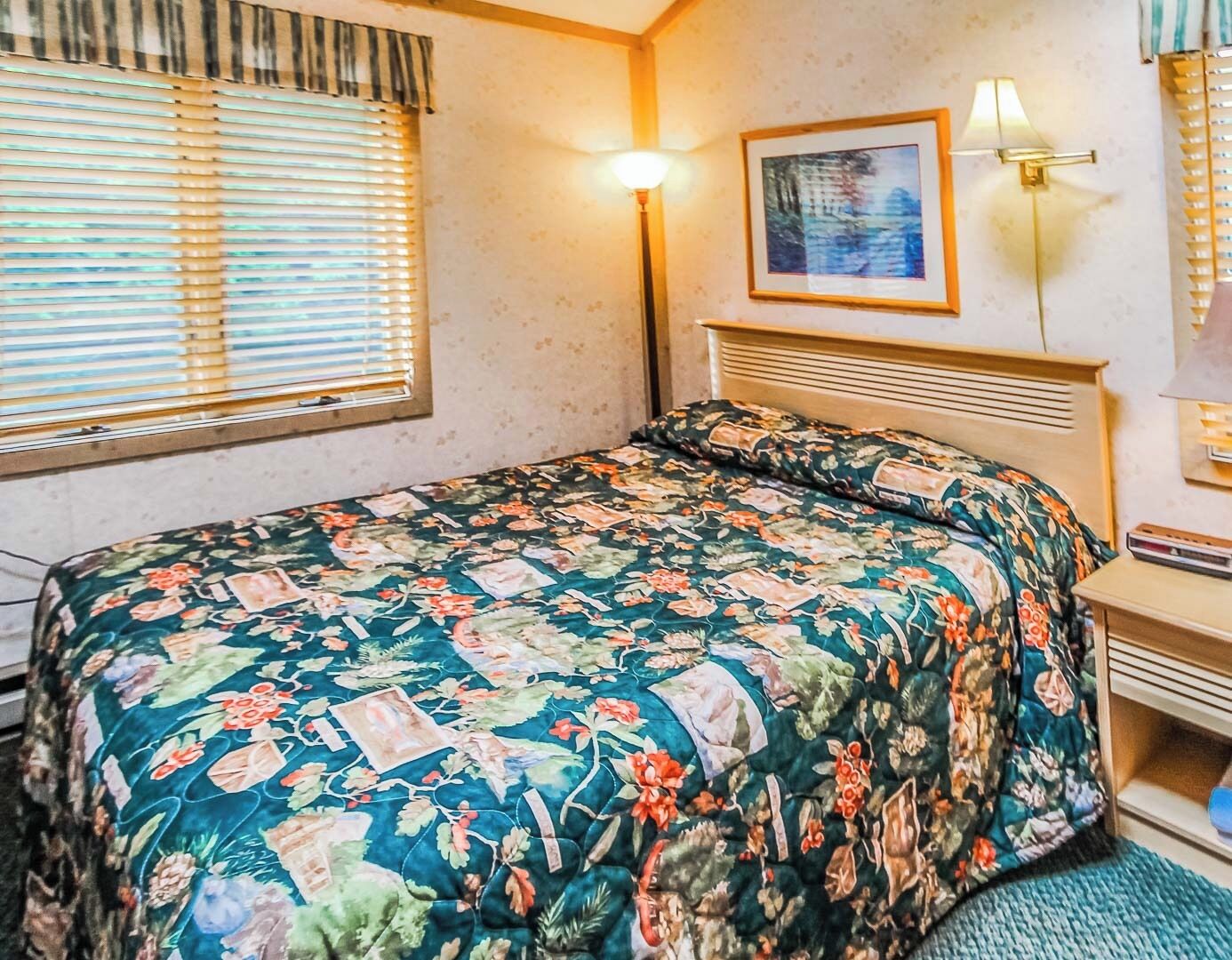 Un dormitorio con colcha estampada, lámpara de noche, un cuadro enmarcado en la pared y una ventana con persianas.