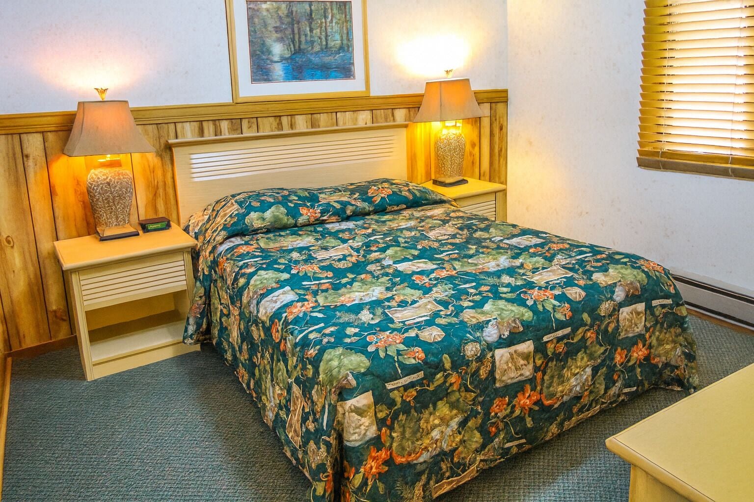 A bedroom with a floral bedspread, wooden headboard, bedside lamps, and a framed picture on the wall.