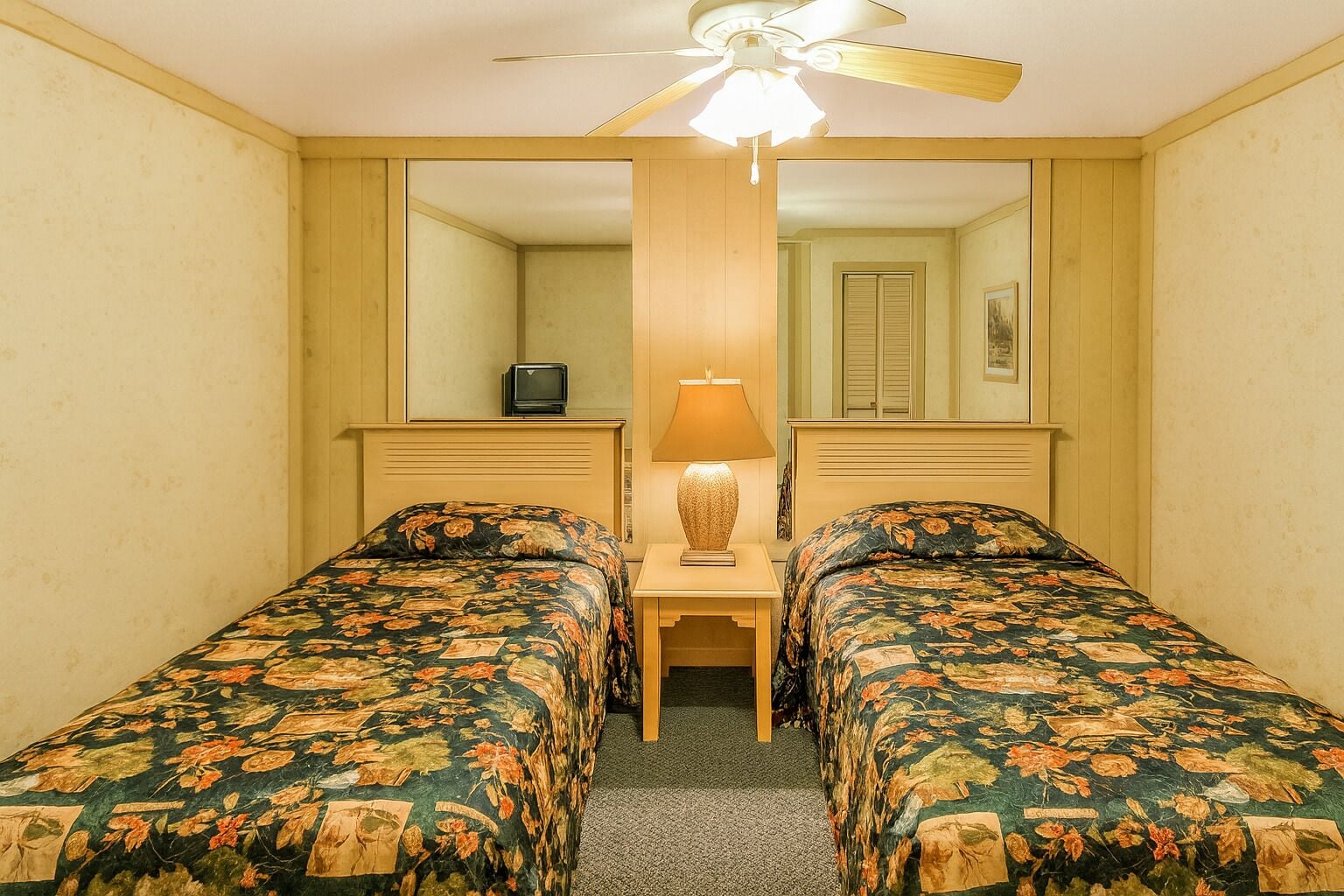 A hotel room with two single beds, a desk with a lamp, a ceiling fan, and a large mirror.
