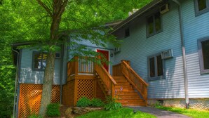 A two-story house with a wooden staircase leading to a red door, surrounded by lush greenery and a stone wall.