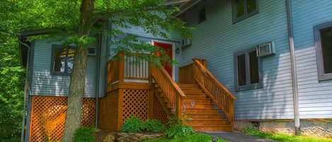 A two-story house with a wooden staircase leading to a red door, surrounded by lush greenery and a stone wall.