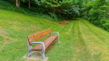 A park bench with metal armrests and a wooden seat, positioned on a grassy slope with a picnic table in the background.