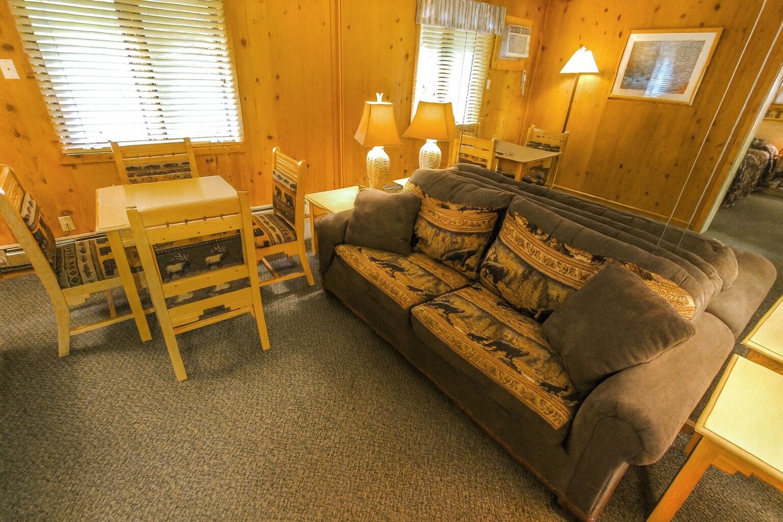 A cozy room with a sofa, a wooden dining table, and chairs. There are two lamps on the table and a framed picture on the wall.