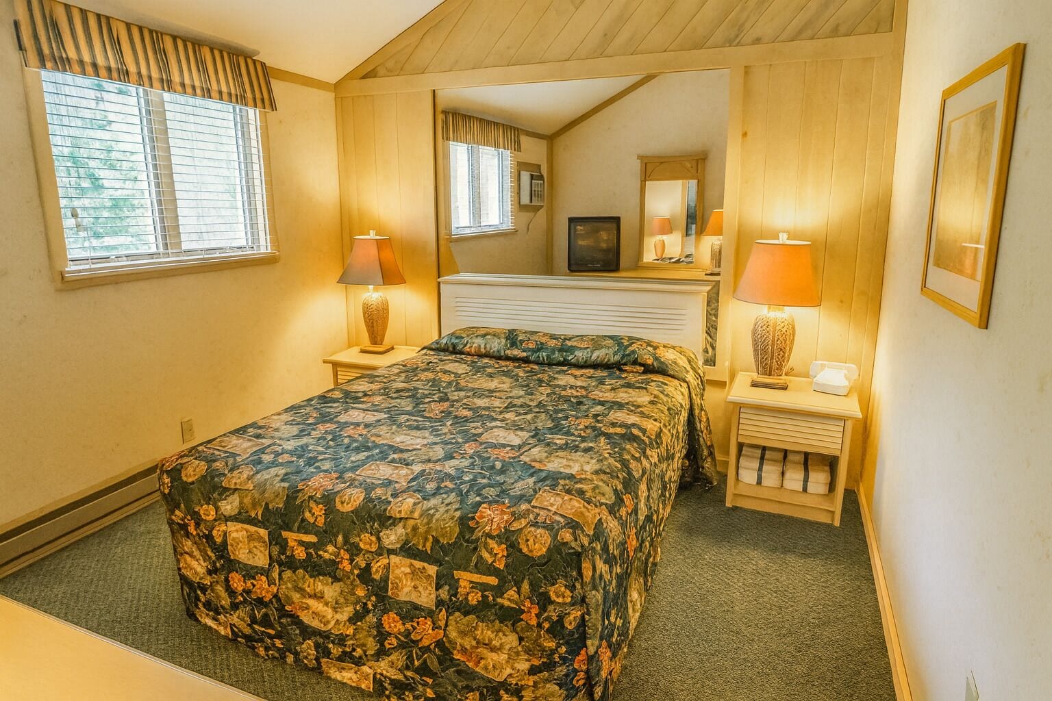 Een slaapkamer met een bed, twee nachtkastjes, lampen, een spiegel en een raam met jaloezieën.
