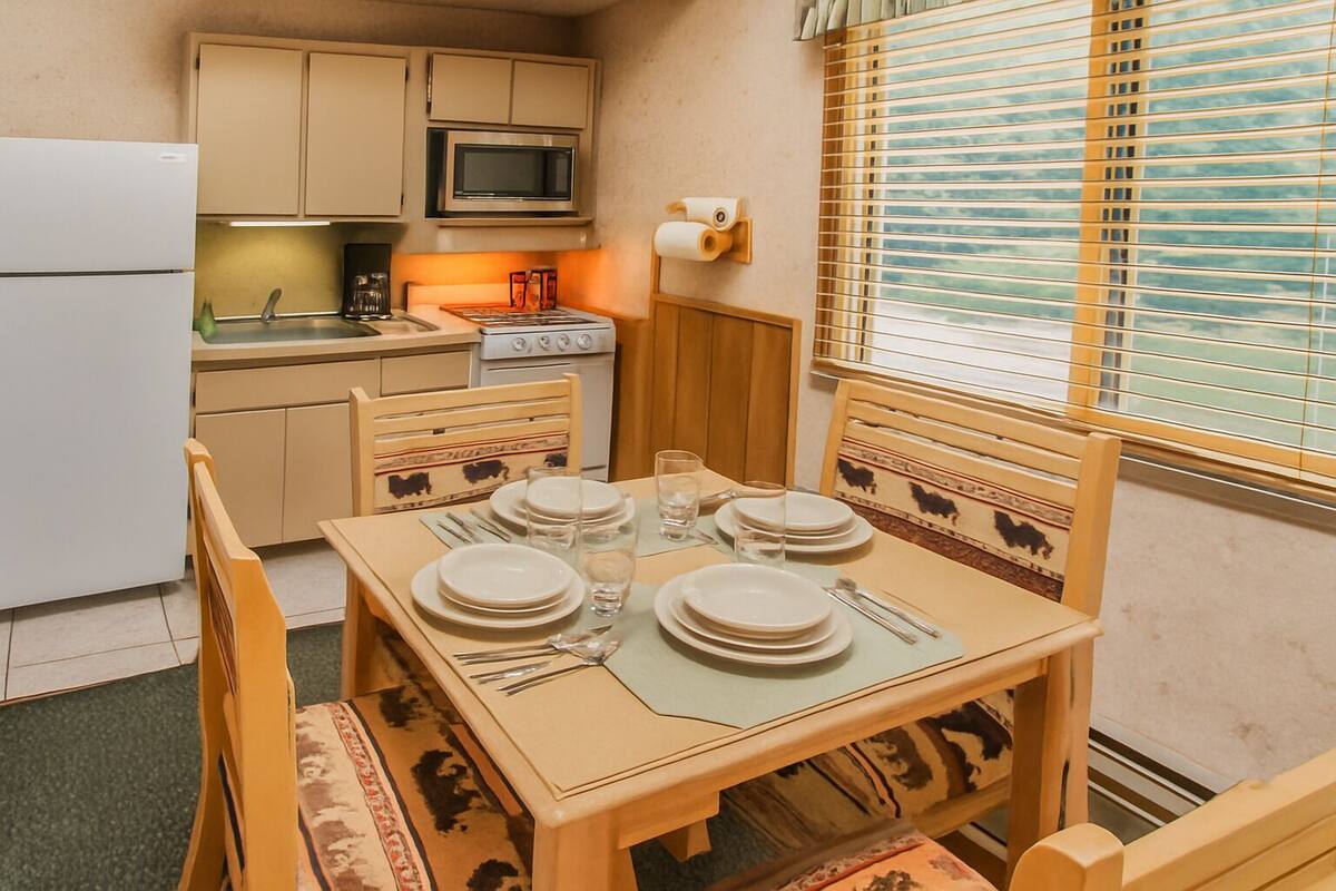 Een kleine keuken met een magnetron, kookplaat en kastjes. Er staat een eettafel gedekt met borden, glazen en bestek.