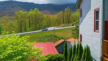 A house with a red roof, a white exterior, and a red window frame. There is a road with a car in the distance, surrounded by green trees and hills.