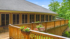 A covered deck with a wooden railing, potted plants, and a view of a house with large windows.