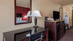 Habitación de hotel con escritorio, silla, televisor, espejo, lámpara y un minibar.