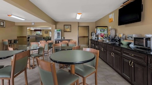 Un comedor de hotel con mesas y sillas redondas, una zona de cocina con microondas, y un televisor montado en la pared.