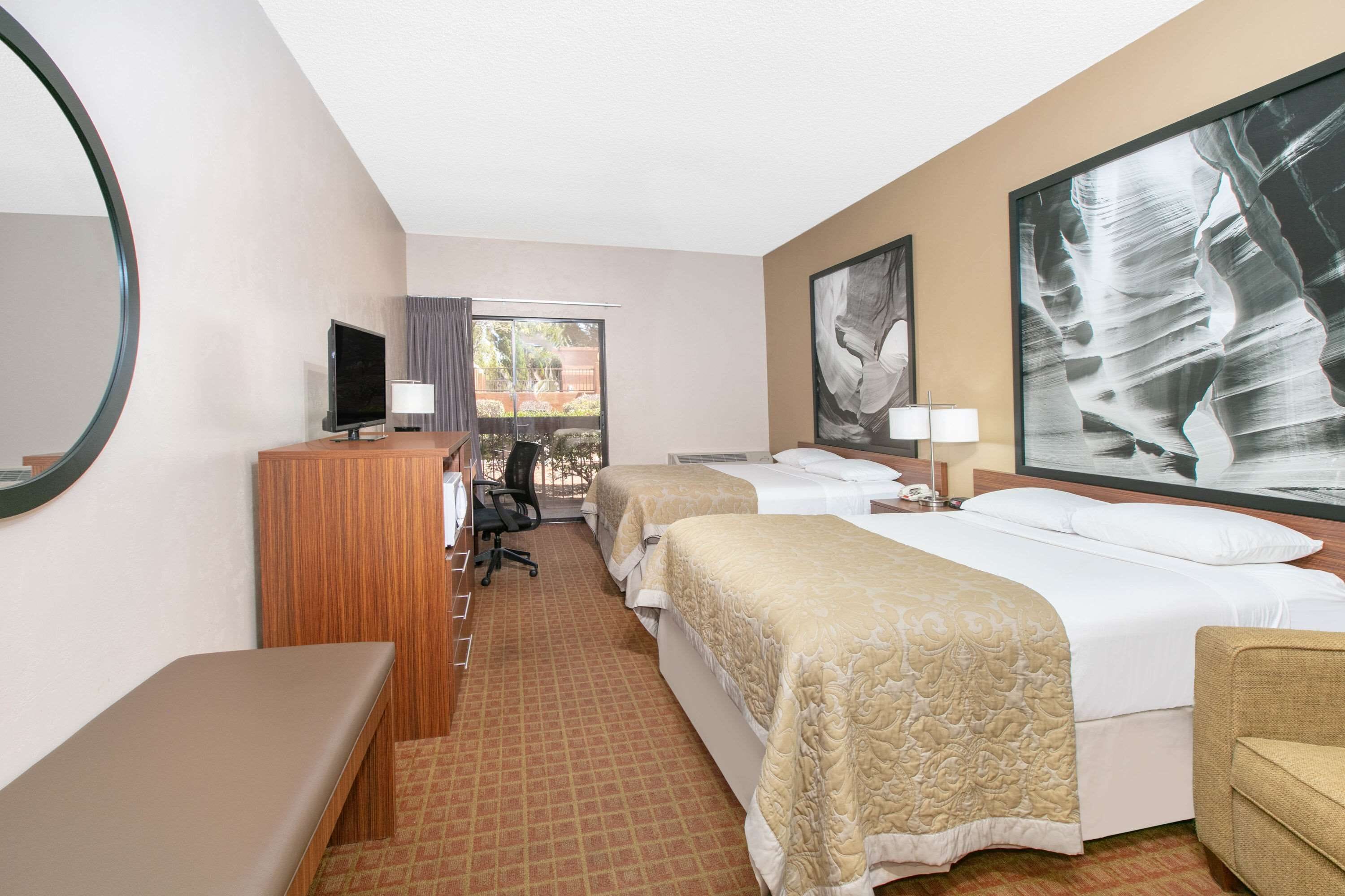 A hotel room with two beds, a desk with a computer, a TV, and a large window with a view of buildings.