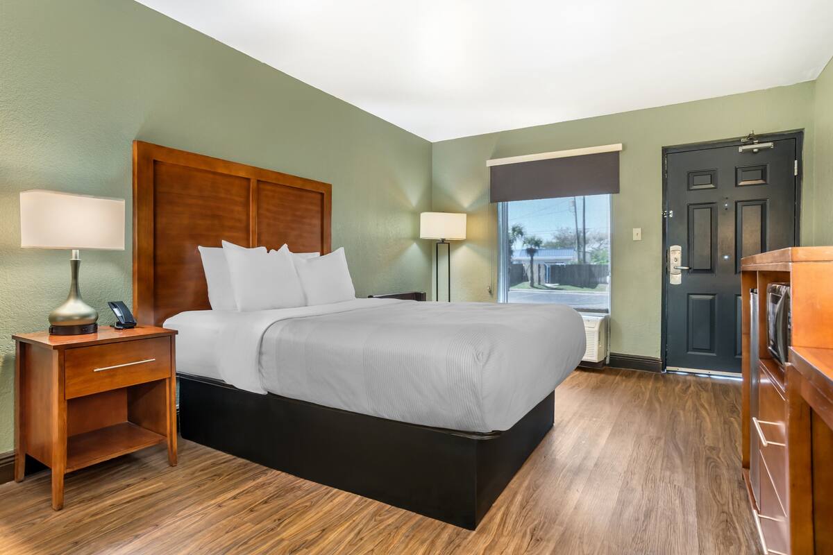 Habitación de hotel con cama, mesitas de noche, lámpara, teléfono, ventana y puerta.