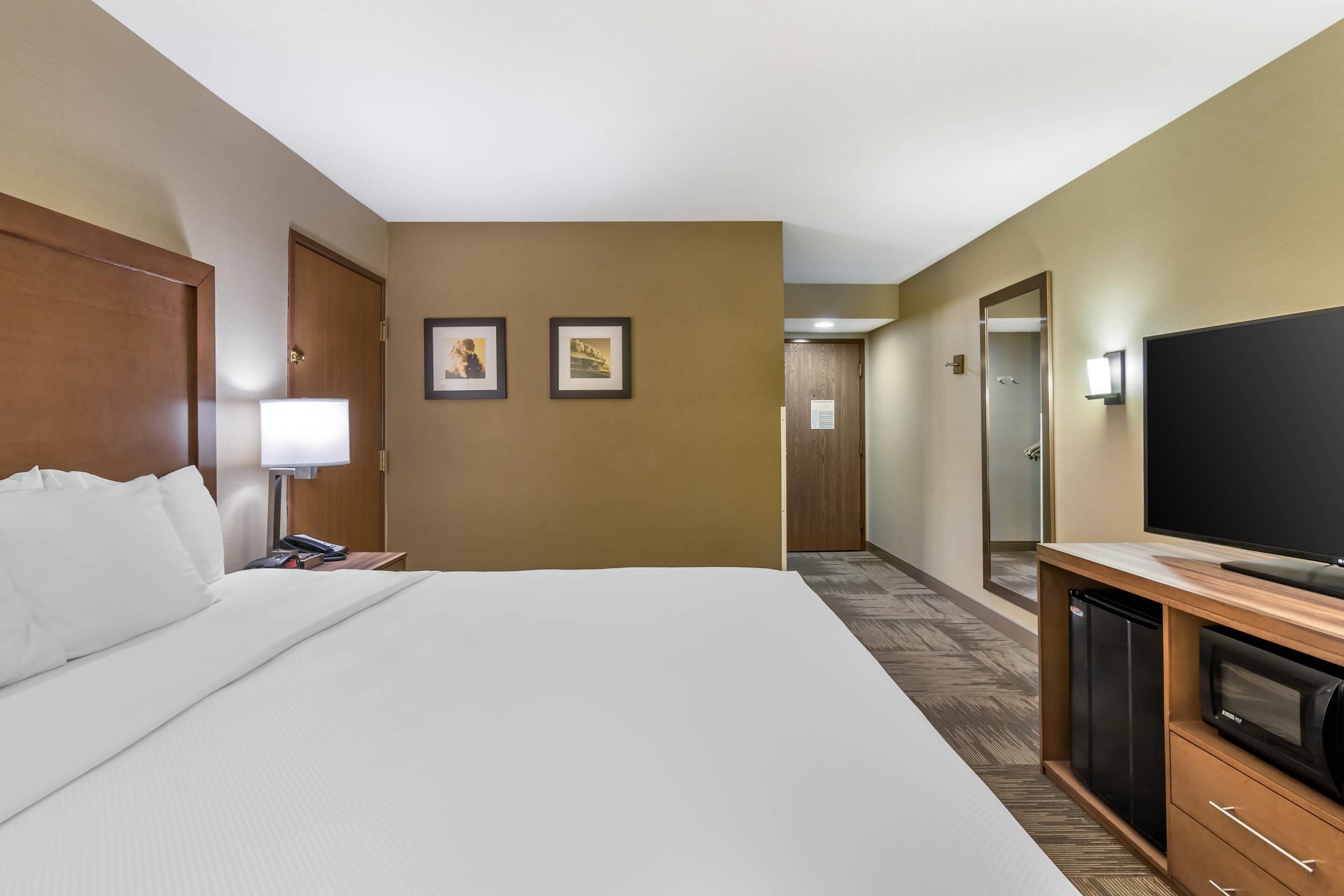 A hotel room with a large bed, a TV, a refrigerator, and a desk with a lamp.