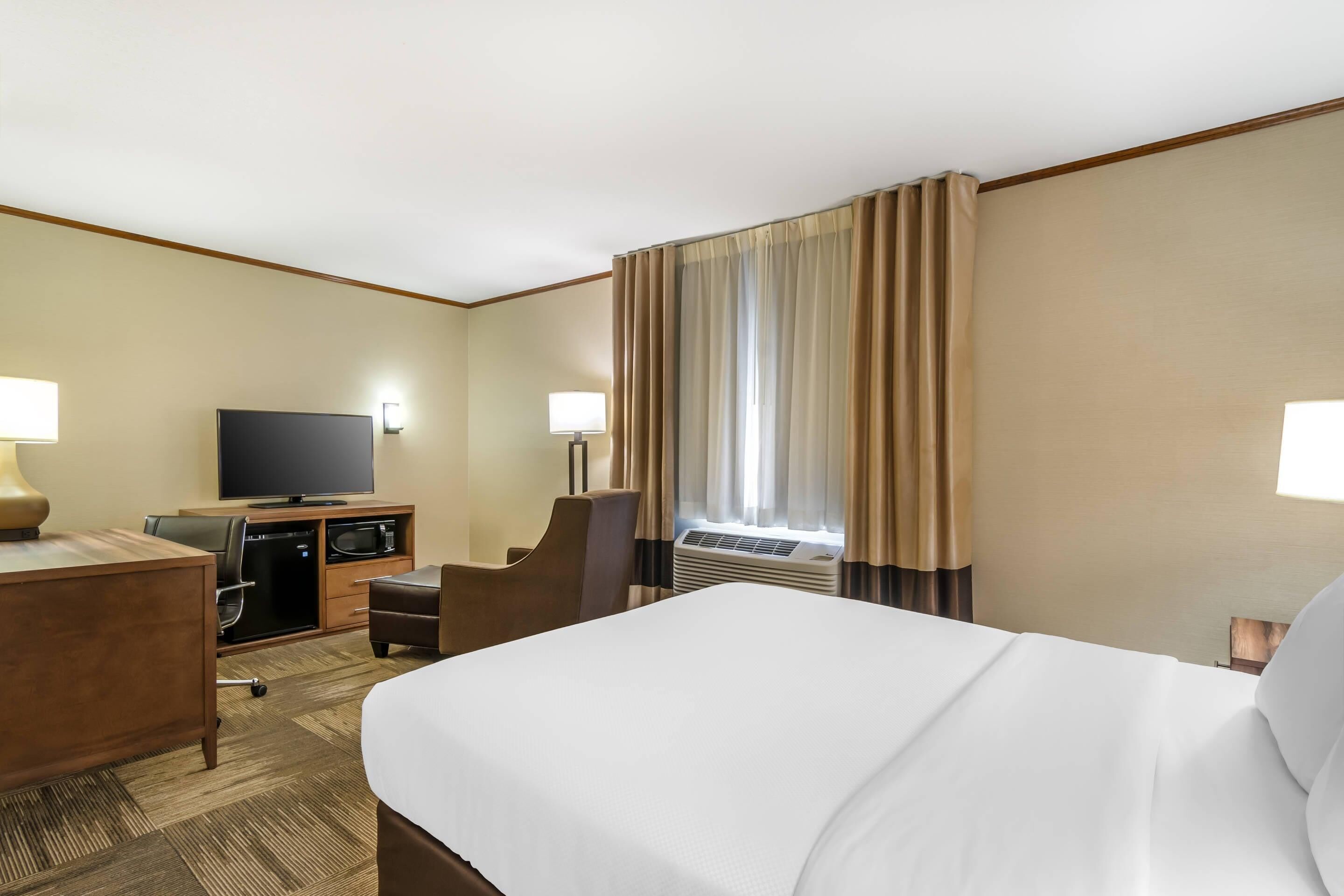 A hotel room with a large bed, a desk with a TV, a chair, a small table, and an air conditioning unit.