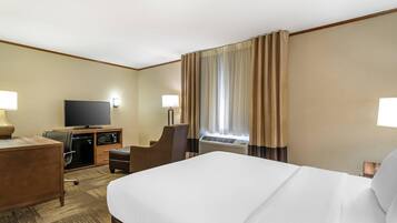 A hotel room with a large bed, a desk with a TV, a chair, a small table, and an air conditioning unit.