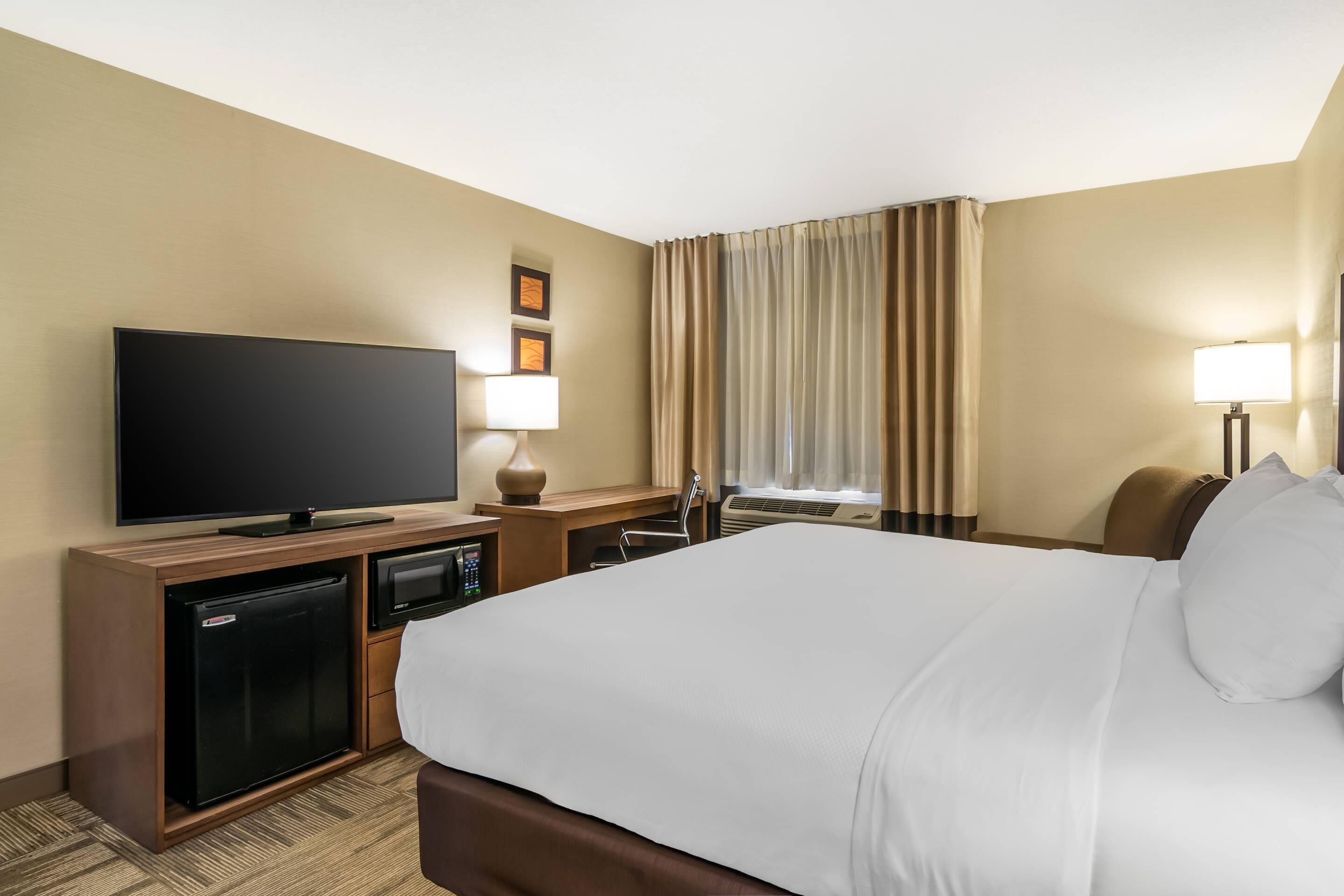 A hotel room with a large bed, a flat-screen TV, a mini-fridge, a desk with a lamp, and a window with curtains.