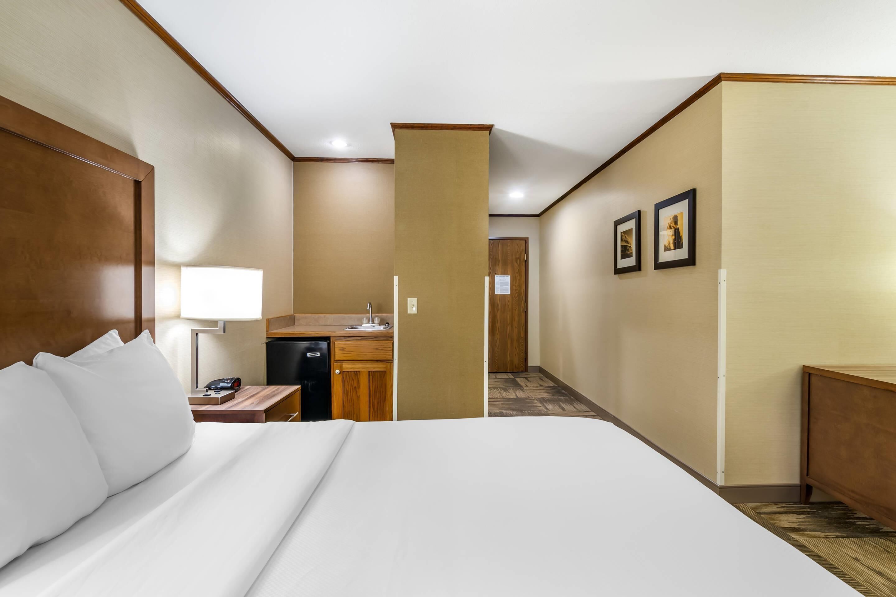Una habitación de hotel con una cama grande, un escritorio con lámpara, una kitchenette pequeña y una puerta que da a otra habitación.