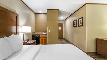 Una habitación de hotel con una cama grande, un escritorio con lámpara, una kitchenette pequeña y una puerta que da a otra habitación.