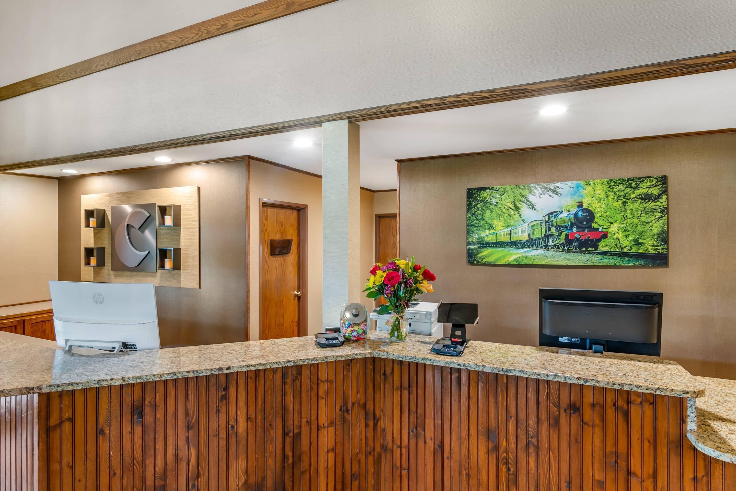 Un espace d’accueil avec un comptoir de réception en bois, un ordinateur de bureau, un téléviseur à écran plat et un tableau représentant un train accroché au mur.
