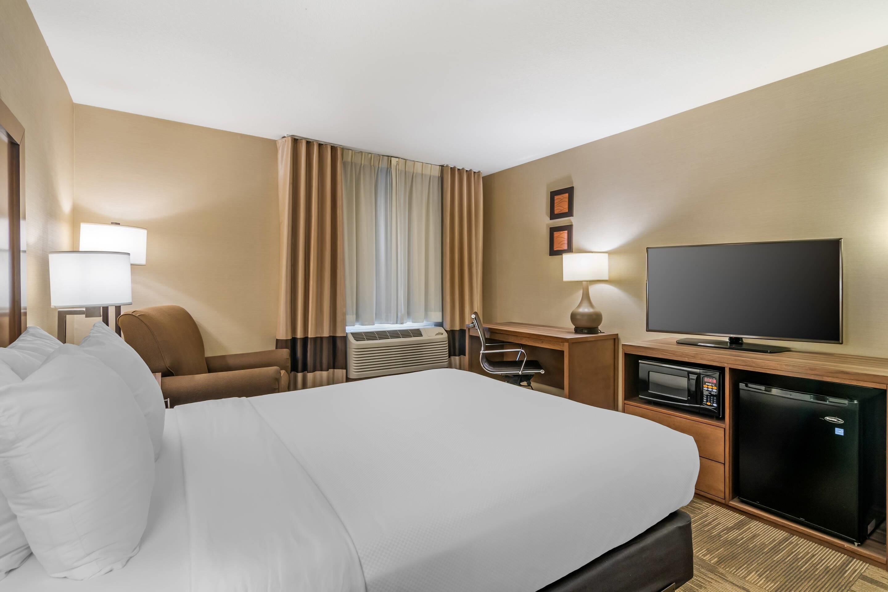 A hotel room with a large bed, a desk with a flat-screen TV, a microwave, a refrigerator, a chair, and a window with curtains.