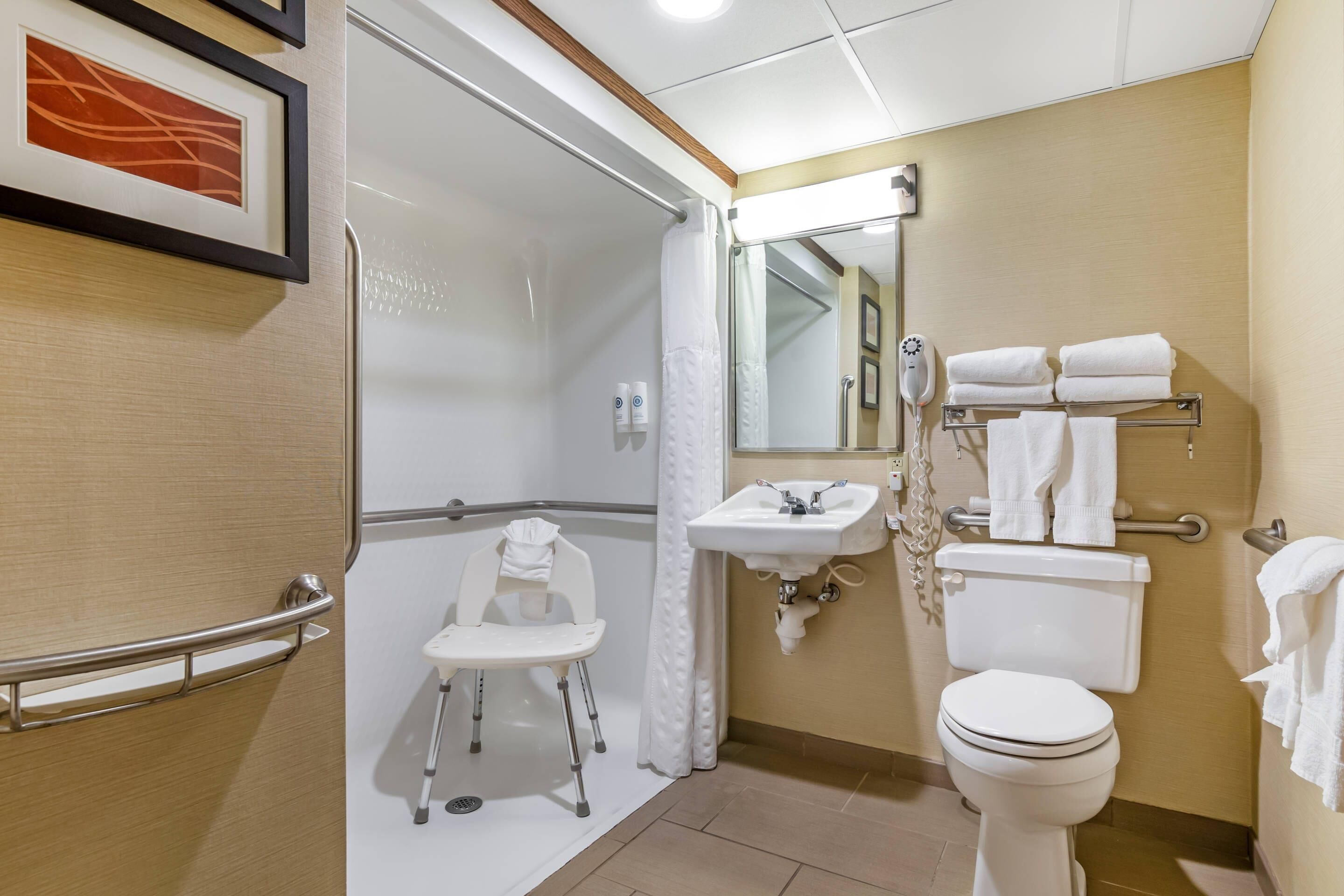A bathroom with a shower, toilet, sink, and a wheelchair-accessible chair.