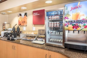 A breakfast bar with a coffee machine, waffle maker, and various food items. There is a juice dispenser and a sign that says "Get your breakfast on."