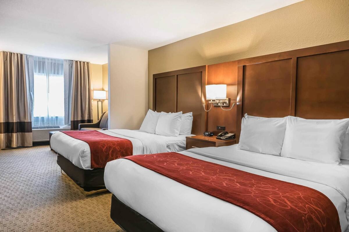 A hotel room with two beds, a wooden headboard, a bedside table with a lamp, and a window with curtains.