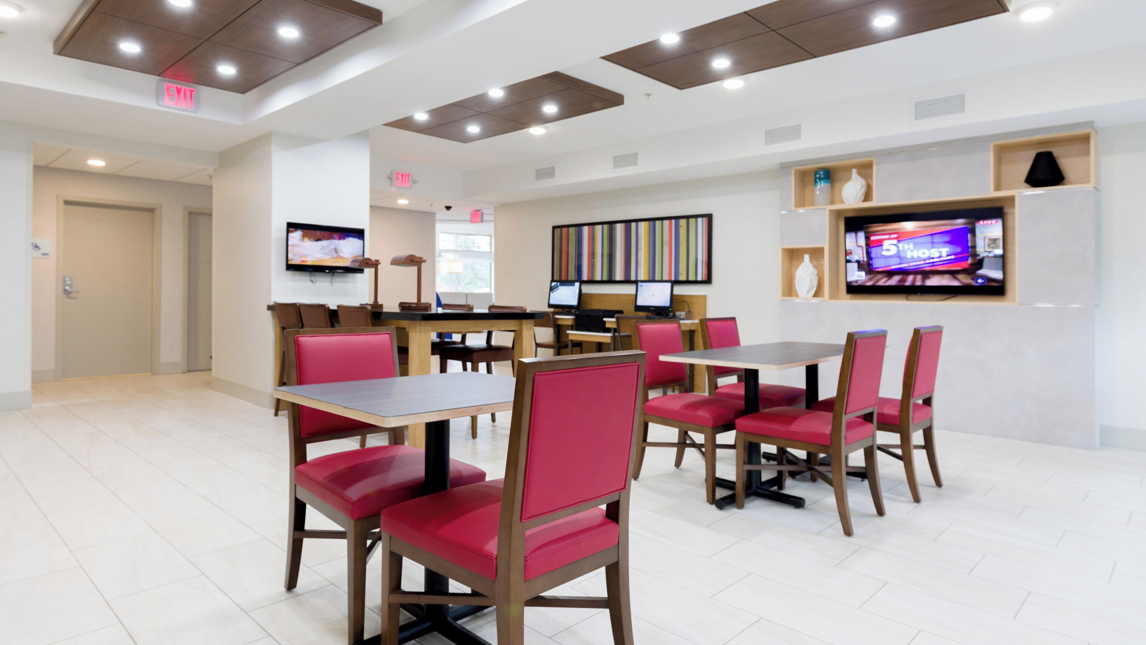 Um restaurante moderno com mesas de madeira e cadeiras estofadas vermelhas, uma televisão de tela plana na parede e um sinal de saída acima da porta.