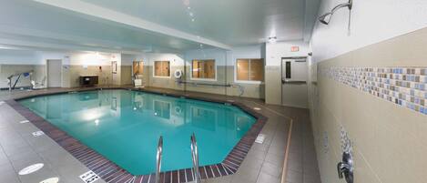 A swimming pool with clear water, surrounded by tiled walls and a tiled floor. There are two lifebuoys and a showerhead visible.