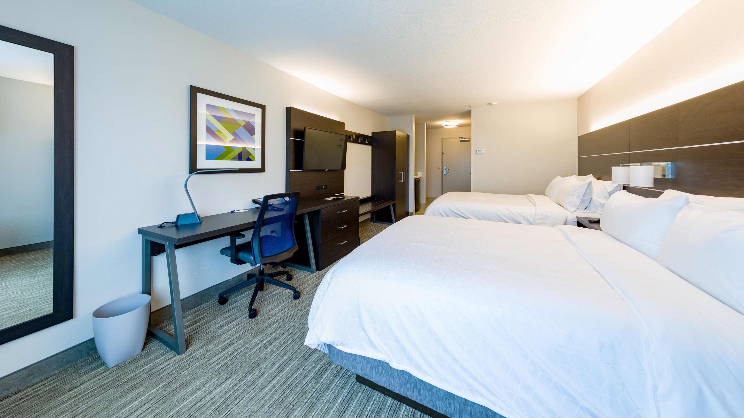A hotel room with a desk, two beds, a TV, and a mirror.