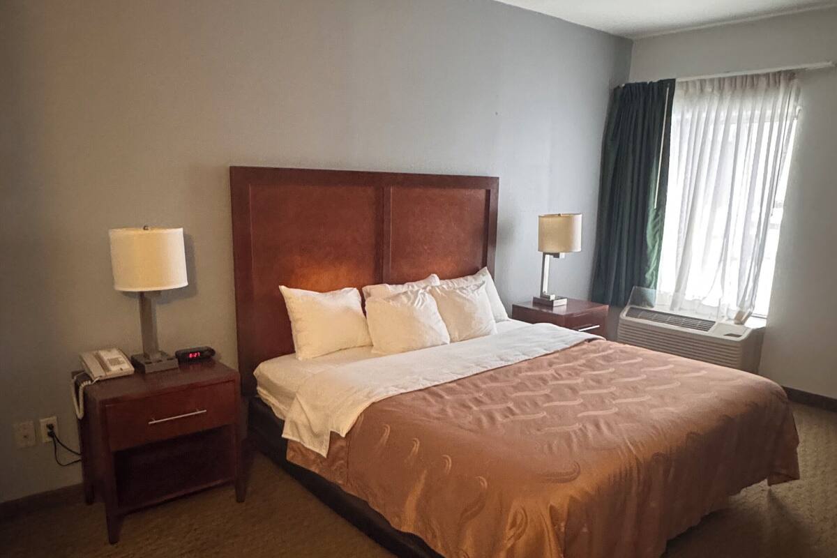 Une chambre d’hôtel avec un grand lit, une table de chevet avec une lampe, une autre table de chevet avec un téléphone et une fenêtre avec des rideaux.