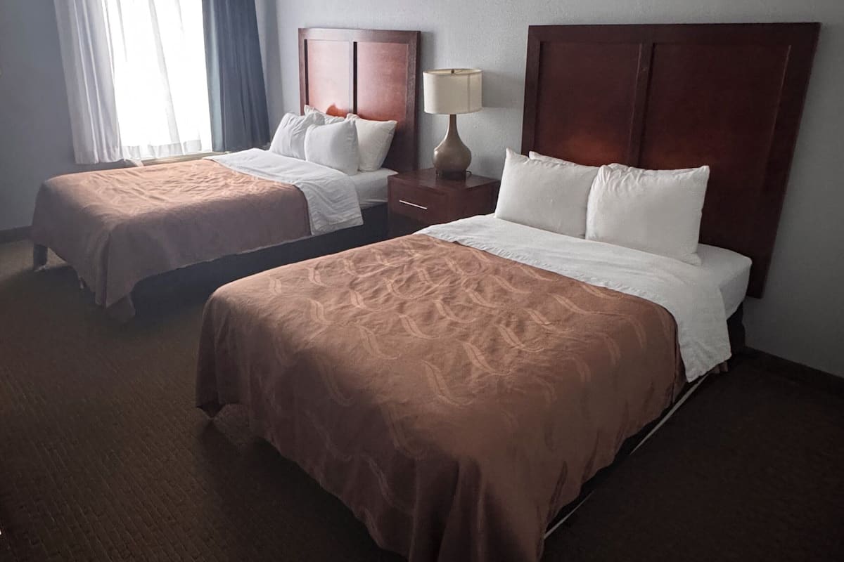 Une chambre d’hôtel avec deux lits, une table de chevet, une lampe et une fenêtre avec des rideaux.