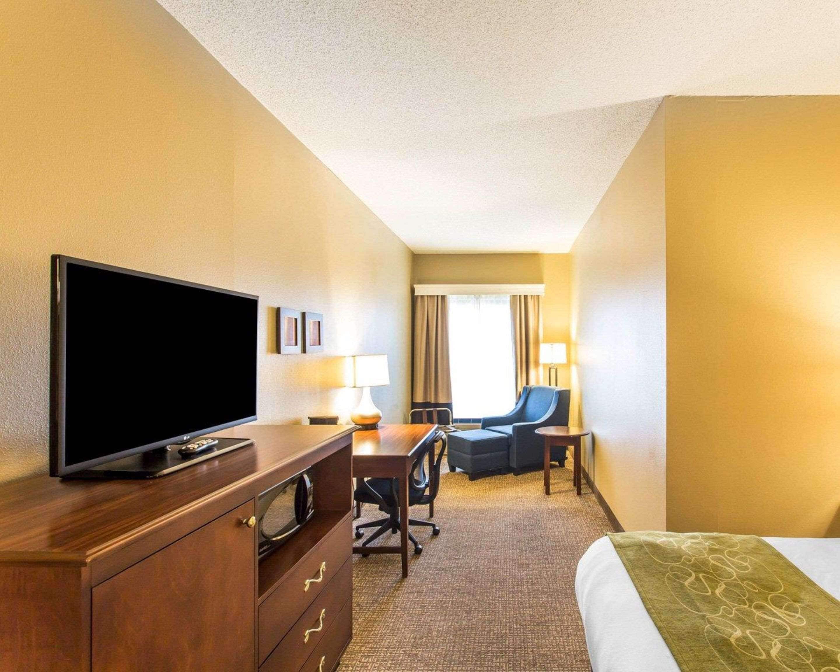 A hotel room with a flat-screen TV, a wooden dresser, a desk with a chair, a bed with a green blanket, and a window with curtains.