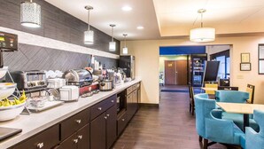A hotel kitchen with a buffet setup, including a coffee station, a refrigerator, and various food items. There are blue chairs and a counter with a laptop.