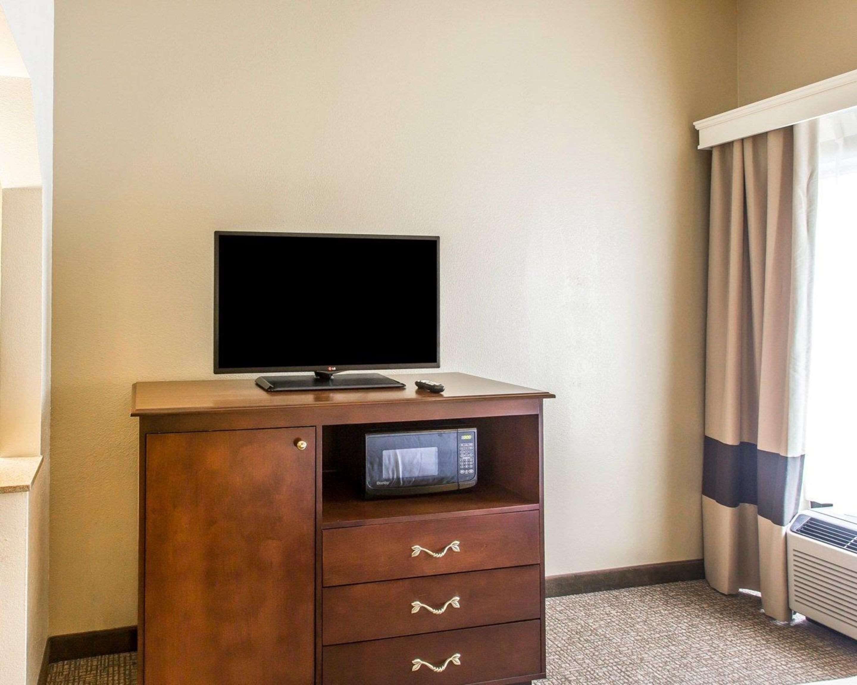 Quarto de hotel com televisão de tela plana sobre um armário de madeira, micro-ondas e janela com cortinas.