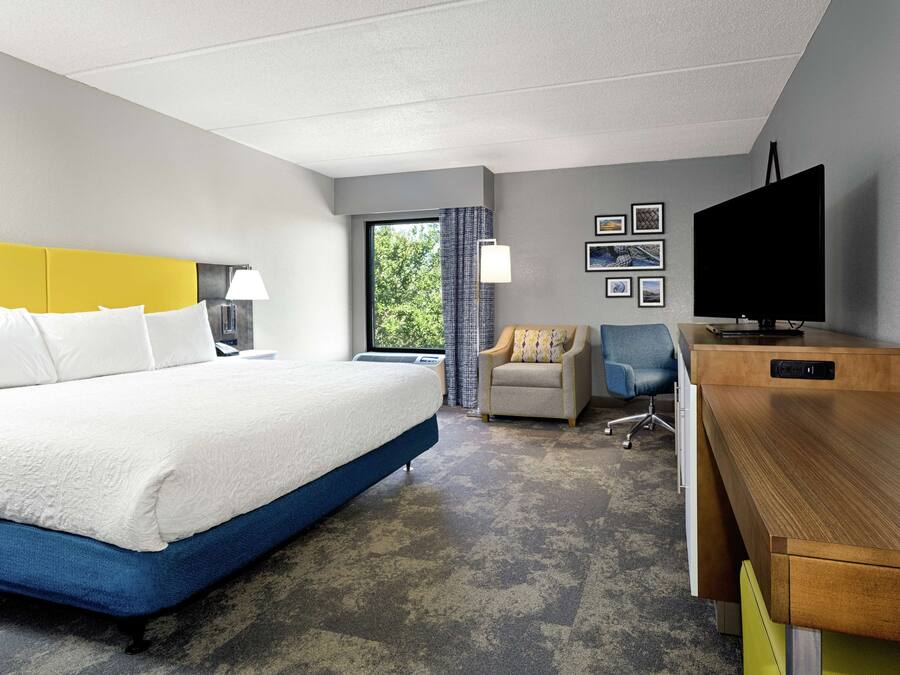 Quarto de hotel com uma cama grande, escrivaninha, cadeira, televisão e janela com cortinas.