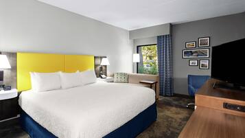 Habitación de hotel con una cama grande, un escritorio, una silla, un televisor y un ventanal con cortinas.