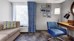 A hotel room with a sofa, a blue armchair, a desk with a lamp, and a window with curtains. - Hampton Inn Amelia Island at Fernandina Beach (Fernandina Beach)