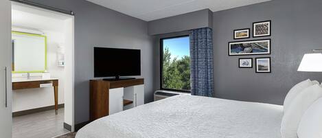 Ein modernes Hotelzimmer mit einem großen Bett, einem Flachbildfernseher, einem Schreibtisch mit Computer und einem Fenster mit Blick auf Bäume.