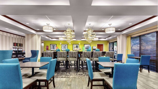 A dining area with multiple tables and chairs, a patterned rug, and a bright yellow accent wall. - Hampton Inn Amelia Island at Fernandina Beach (Fernandina Beach)