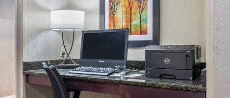 A hotel reception desk with a desktop computer, a printer, a framed picture on the wall, and a desk lamp.