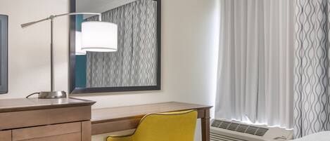 A hotel room with a wooden desk, a yellow chair, a large mirror, and a window with patterned curtains.