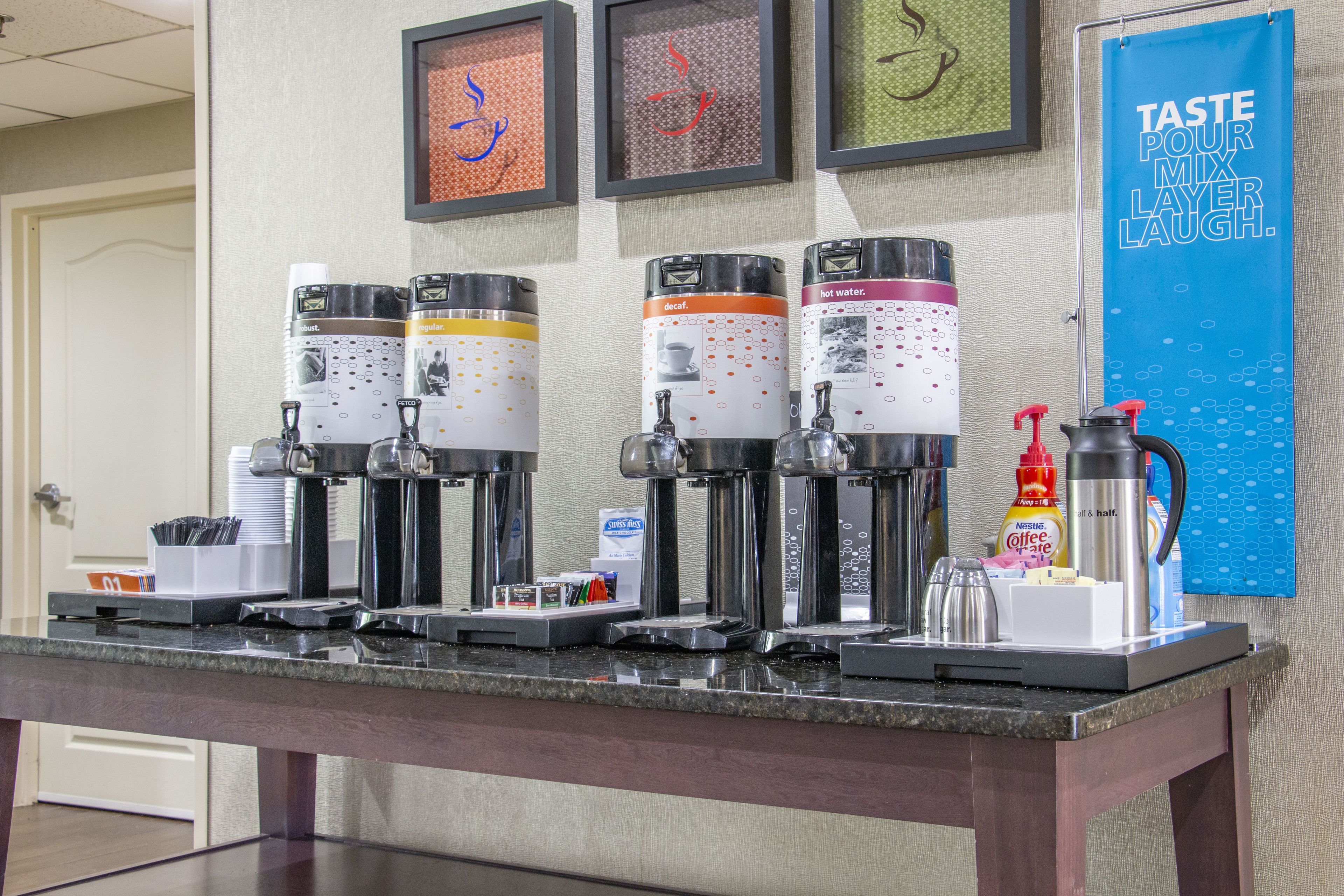 Una estación de café con varios dispensadores, un cartel que dice "TASTE POUR MIX LAYER LAUGH." y una obra de arte enmarcada en la pared.