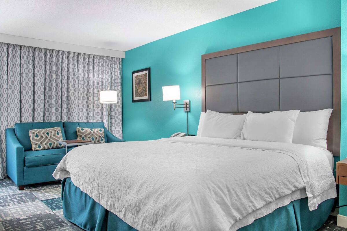 A hotel room with a large bed, a blue armchair, and a patterned curtain.