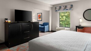 Ein Hotelzimmer mit einem großen Bett, einem Fernseher auf einem Schrank, einem Sessel, einem Schreibtisch mit Lampe und einem Fenster mit Blick ins Grüne.