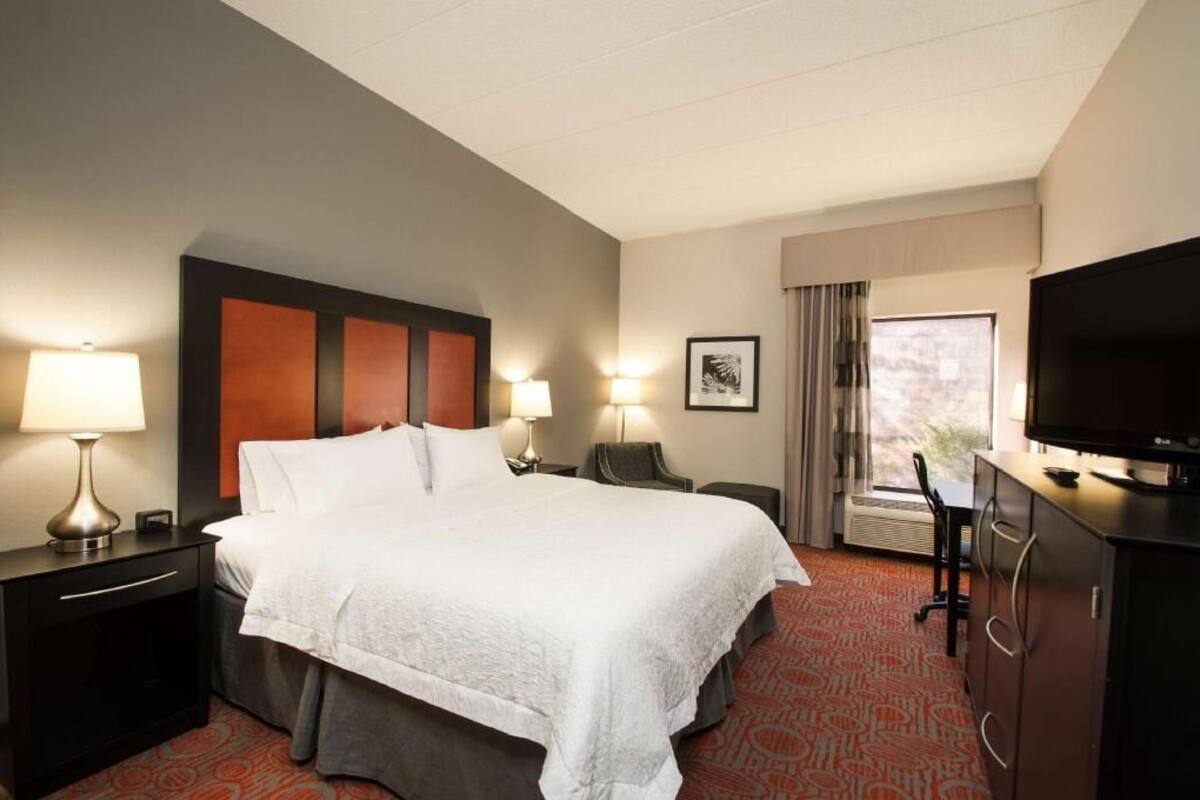 A hotel room with a large bed, two bedside lamps, a desk with a chair, a television, and a window with curtains.
