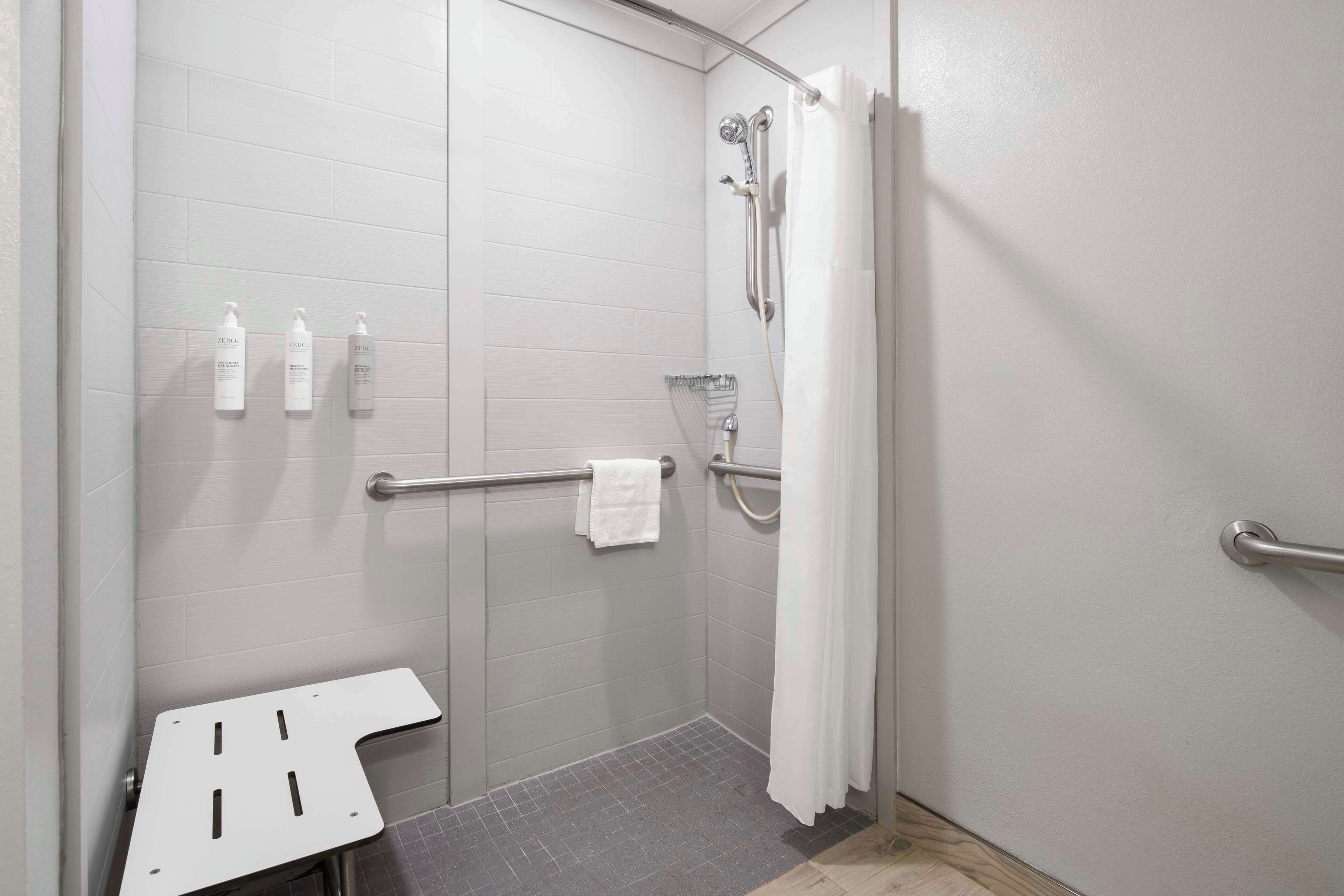 Ein modernes Badezimmer mit barrierefreier Dusche, Haltegriffen, einem weißen Handtuch und Pflegeprodukten an der Wand.