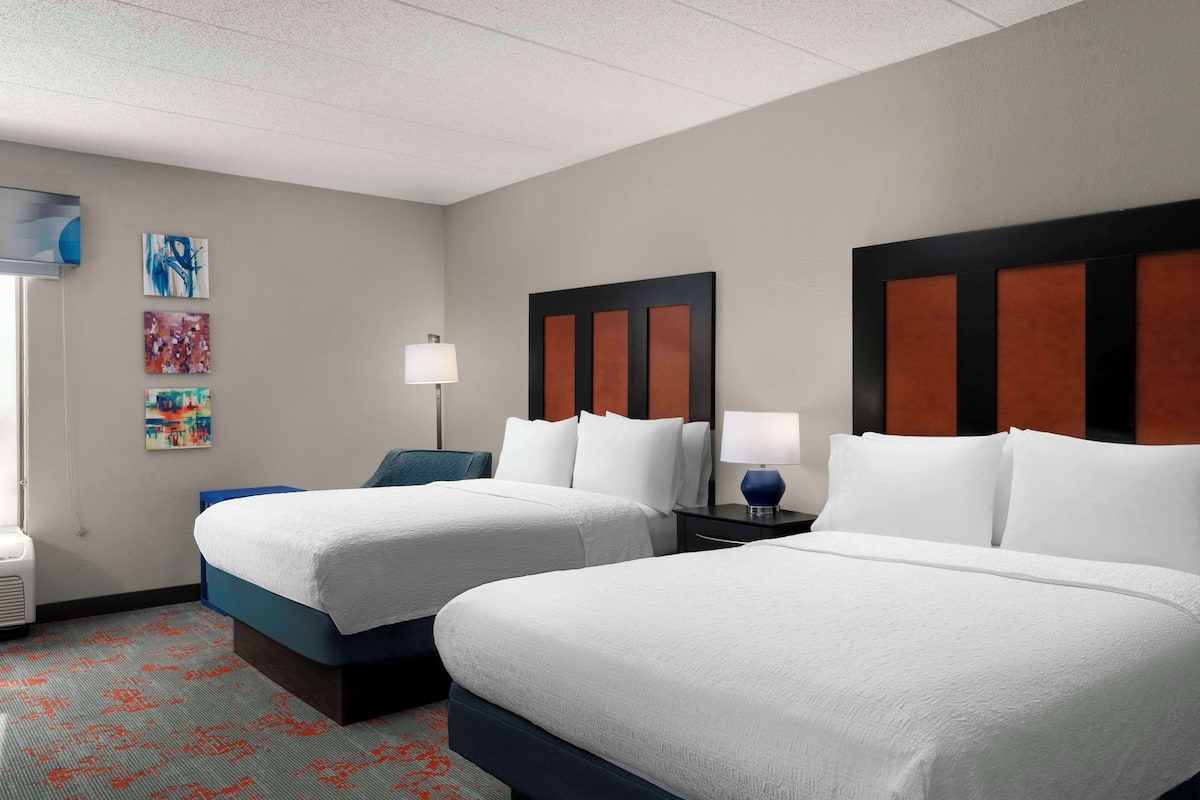 A hotel room with two beds, a desk, a chair, and artwork on the wall.