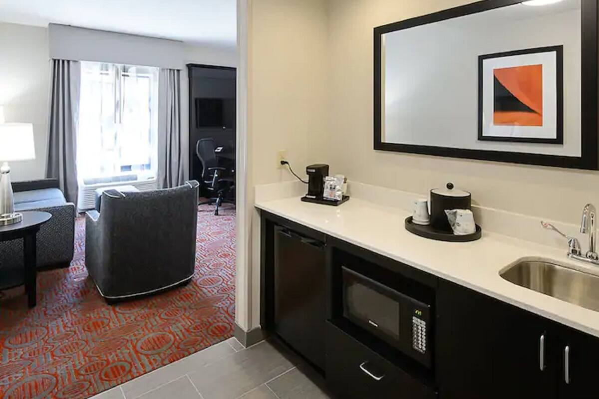 A hotel room with a kitchenette, a dining area with a chair and table, a large window with curtains, and a framed picture on the wall.