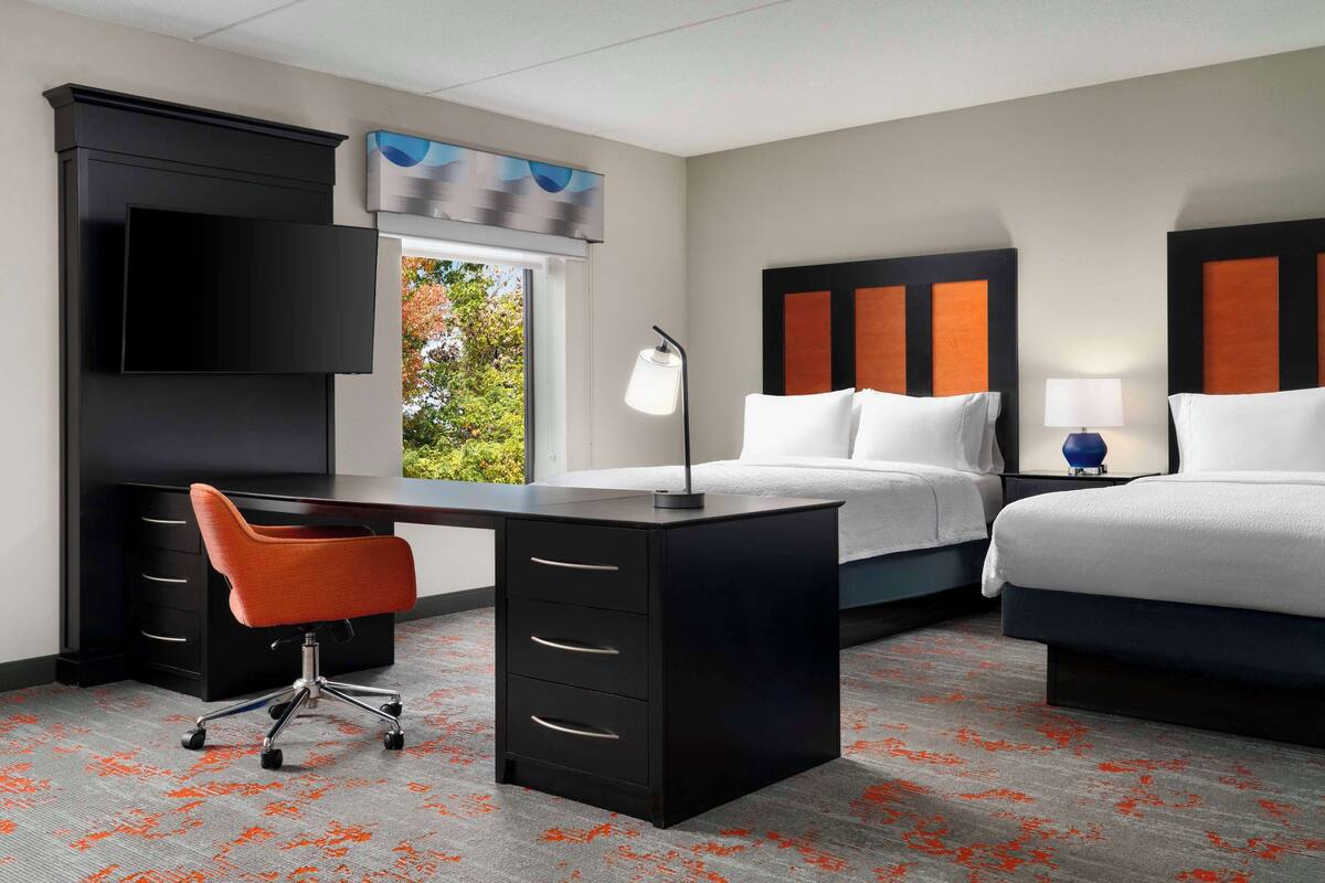 A hotel room with a desk, two beds, a chair, and a window with a view of greenery.