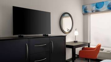 A hotel room with a flat-screen TV on a black cabinet, a round mirror on the wall, a desk with a lamp, an orange chair, and a large window with a view.